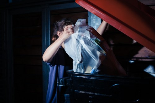 A person with short, dark hair is seen disposing of a clear plastic bag filled with assorted rubbish into a black waste bin. The individual is wearing a dark blue shirt and is standing next to a large outdoor waste container. The scene is set against a background of wooden paneling with visible blue framework, likely part of a garden or outdoor storage area. The waste bin is positioned on a paved surface, and a red lid is partially lifted by the person as they deposit the rubbish. The lighting appears artificial, casting shadows and highlighting the plastic bag, which contains mixed waste materials. This image reflects typical activities associated with rubbish removal services, such as those offered by Garden Clearance Pimlico, who provide garden clearance and waste disposal in the Pimlico area, ensuring proper and professional rubbish collection.