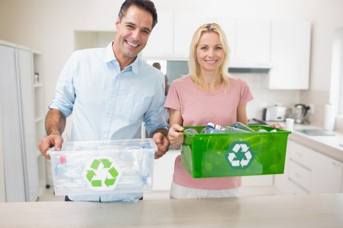 Two smiling individuals, a man and a woman, standing in a modern kitchen with white cabinets and appliances in the background, each holding a recycling bin with a green recycling symbol on the front. The man, dressed in a light blue shirt, holds a transparent plastic bin filled with clear bottles and packaging, while the woman, wearing a pink top, holds a solid green bin with similar recycling branding. They are positioned in the foreground on a light-colored kitchen counter, and the environment appears clean, well-lit, and organized, suggesting a focus on waste sorting and proper rubbish management. This scene reflects responsible disposal practices, aligning with rubbish removal and recycling services such as those offered by Garden Clearance Pimlico for domestic properties in the Pimlico area.
