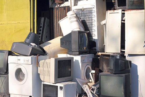 A collection of discarded household appliances and electronic waste arranged outdoors on a paved surface, including white washing machines, old computer monitors, and a microwave oven, with some items leaning against a metal shed or storage area. The appliances vary in size and condition, featuring visible glass screens, plastic casings, and metallic components with minor signs of dirt and weathering. The background includes parts of a metal storage structure with corrugated panels and a yellow wall, indicating a waste clearance or recycling collection point, typical of services offered by Garden Clearance Pimlico to remove unwanted household rubbish in the local Pimlico area. The scene is illuminated by natural daylight, casting soft shadows on the items, highlighting the textures of plastic, glass, and metal surfaces, and emphasizing the cluttered nature of typical household rubbish accumulation for removal or disposal.